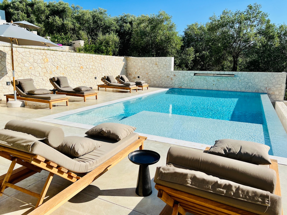 A serene outdoor area is presented, featuring a crystal-clear infinity pool surrounded by olive trees. Several loungers with soft cushions are positioned near the pool, creating inviting spots for relaxation. A round side table stands between the loungers, enhancing the tranquil setting.