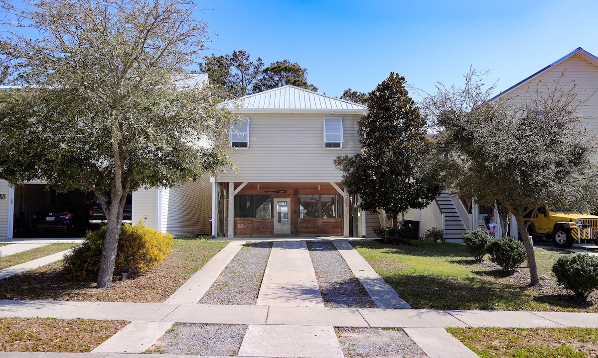 A two-story beach home is positioned among landscaped greenery and manicured lawns. The building features light-colored siding and a metallic roof, with large glass entry doors welcoming visitors. Sidewalks lead towards the entrance, framed by trees and shrubs, providing a sense of openness.