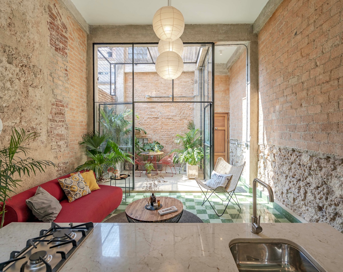 A cozy living area features a red sofa adorned with decorative pillows, complemented by a round coffee table. Large glass doors provide a view of a lush interior patio filled with tropical plants. Natural light floods the space through the high ceiling, accentuating the exposed brick walls.