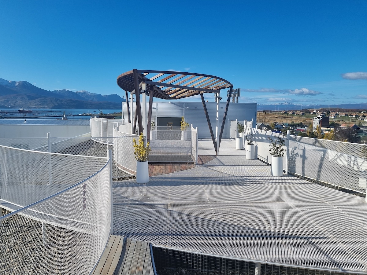 The rooftop terrace offers a spacious area with panoramic views of the city and surrounding mountains. A wooden structure provides shade, while planters with greenery enhance the open space. The terrace features a seamless mesh railing, promoting safety without obstructing the view.