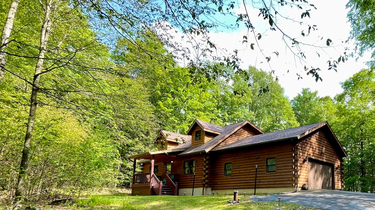 The exterior of a log cabin is framed by lush greenery, featuring a front porch and a two-car garage. Sunlight filters through the leaves, highlighting the natural wood finish of the cabin. A serene, wooded area surrounds the property, enhancing its secluded feel.