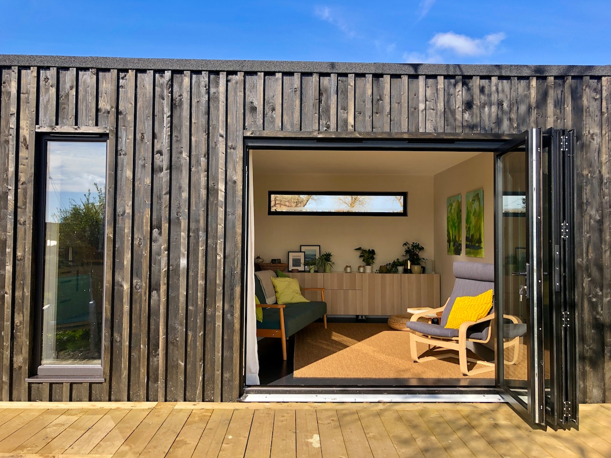 A modern detached building with a wooden exterior is depicted, featuring large glass doors that open to reveal a light-filled living area. Inside, comfortable seating is complemented by greenery and artistic accents, while a serene outdoor deck extends the living space.