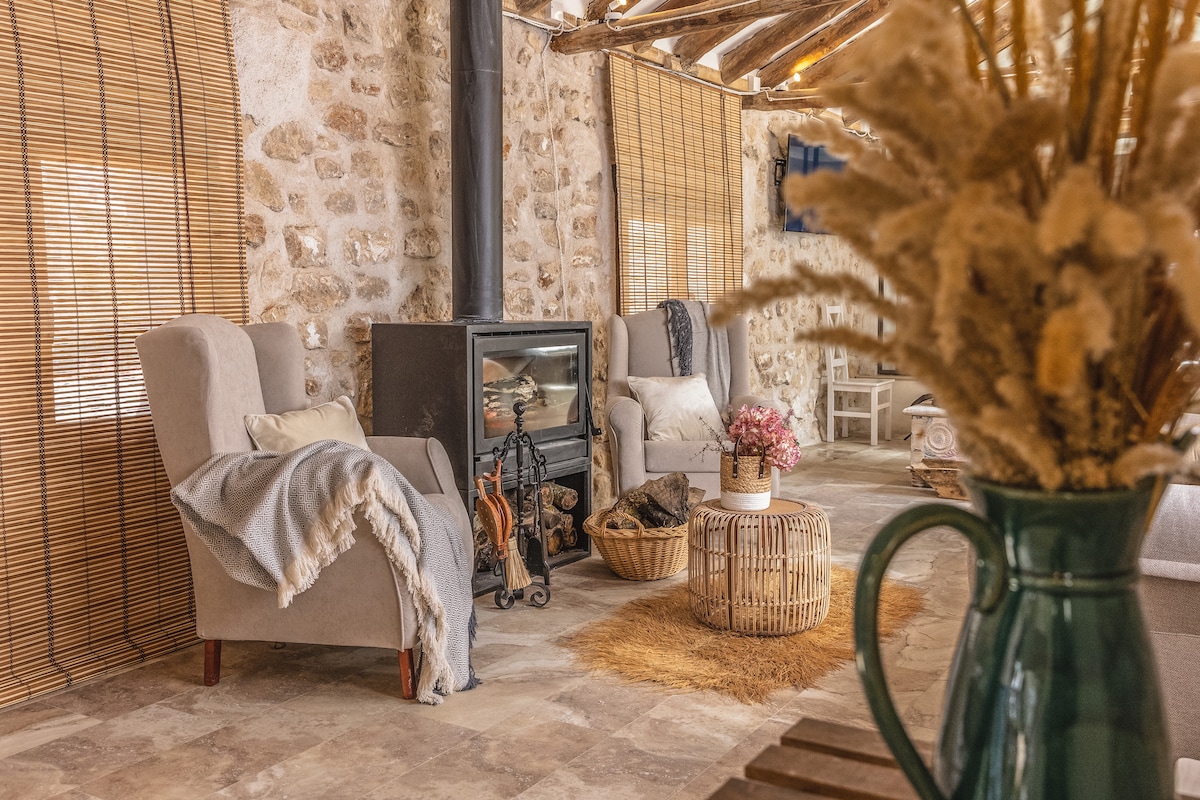 The inviting living space features two armchairs positioned near a wood-burning stove. A soft blanket drapes over one chair, and a woven basket holds a cozy throw. A rustic table showcases a bouquet of dried flowers, adding warmth to the stone walls and ceiling beams.