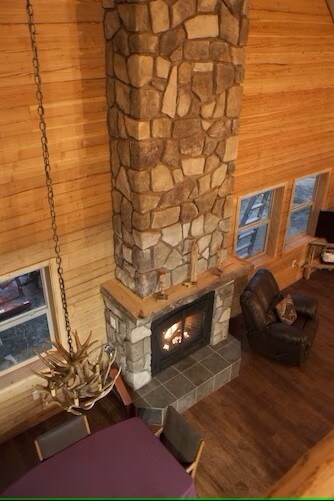 A stone fireplace is centrally positioned within the lodge, featuring a wood-burning fire. A leather armchair is placed nearby, complemented by warm wood paneling on the walls. Large windows allow natural light to flow in, illuminating the cozy living space.