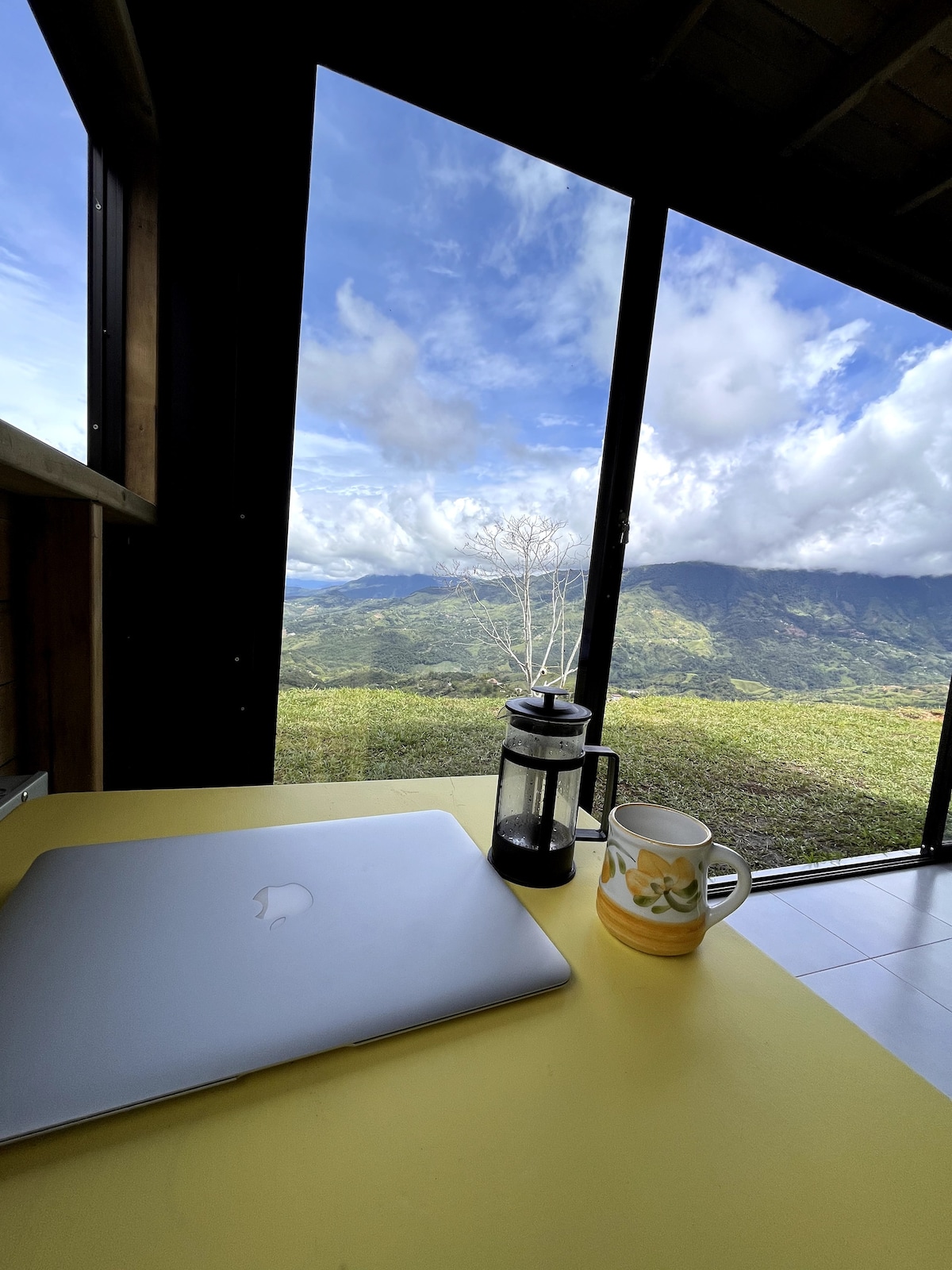 A workspace is positioned near a large window, revealing expansive views of lush green mountains under a partly cloudy sky. A laptop, a French press, and a mug are placed on a bright yellow table, inviting a productive atmosphere.