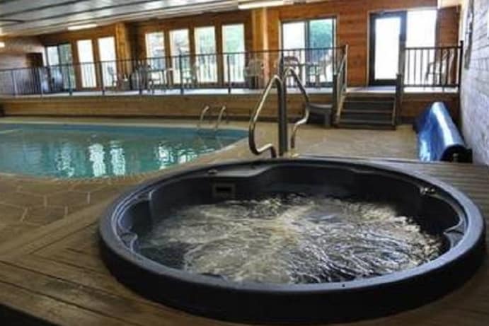 An indoor pool area is visible, featuring a hot tub in the foreground. The pool has clear water and is surrounded by wooden decking. Large windows allow natural light to fill the space, providing views of the outside. Chairs are positioned by the poolside.