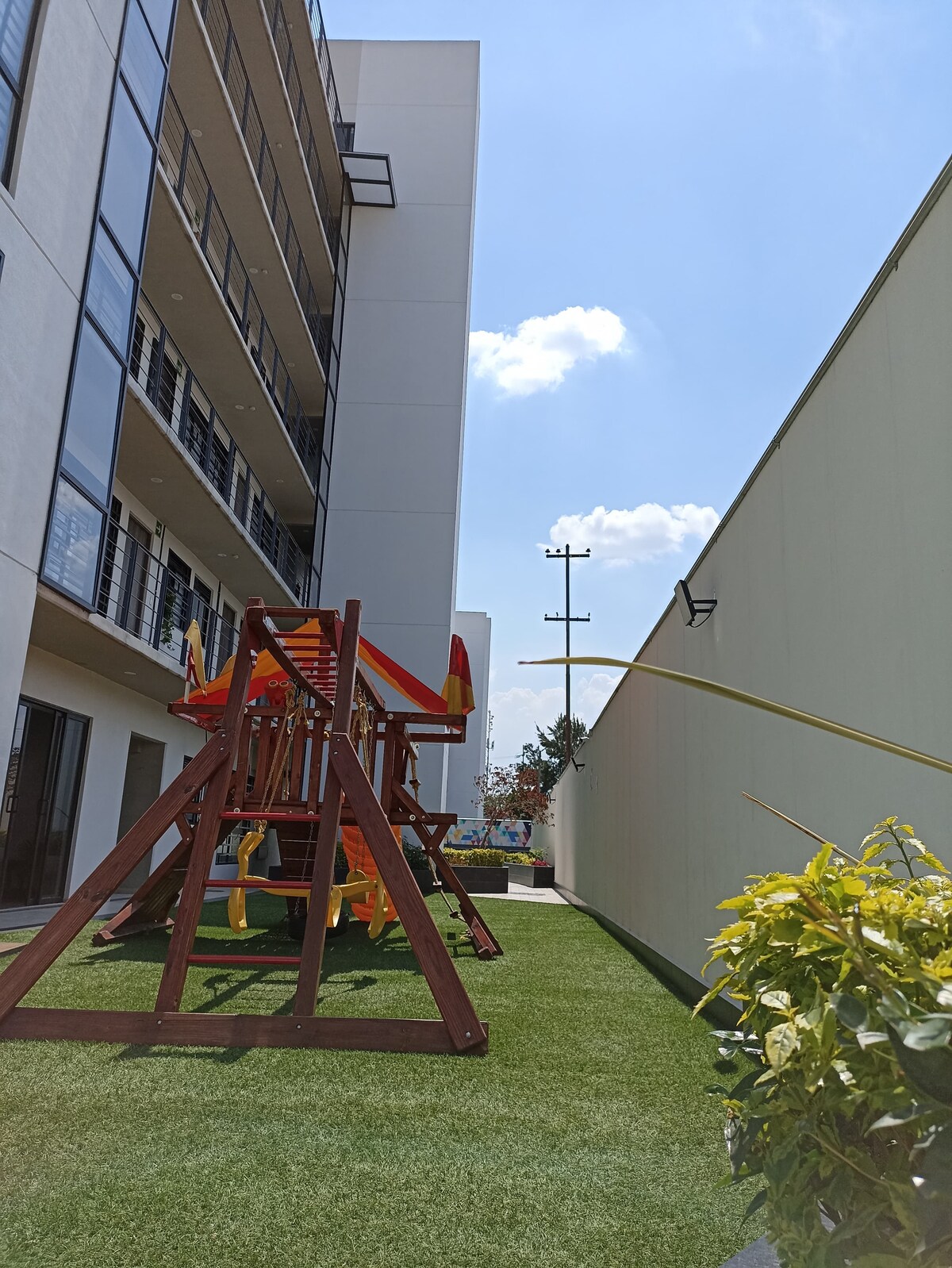 A playground area features a wooden playset with slides and swings, positioned on artificial grass. The bright sunny sky complements the outdoor space, framed by a modern apartment building and a wall, suggesting a secure and inviting environment.