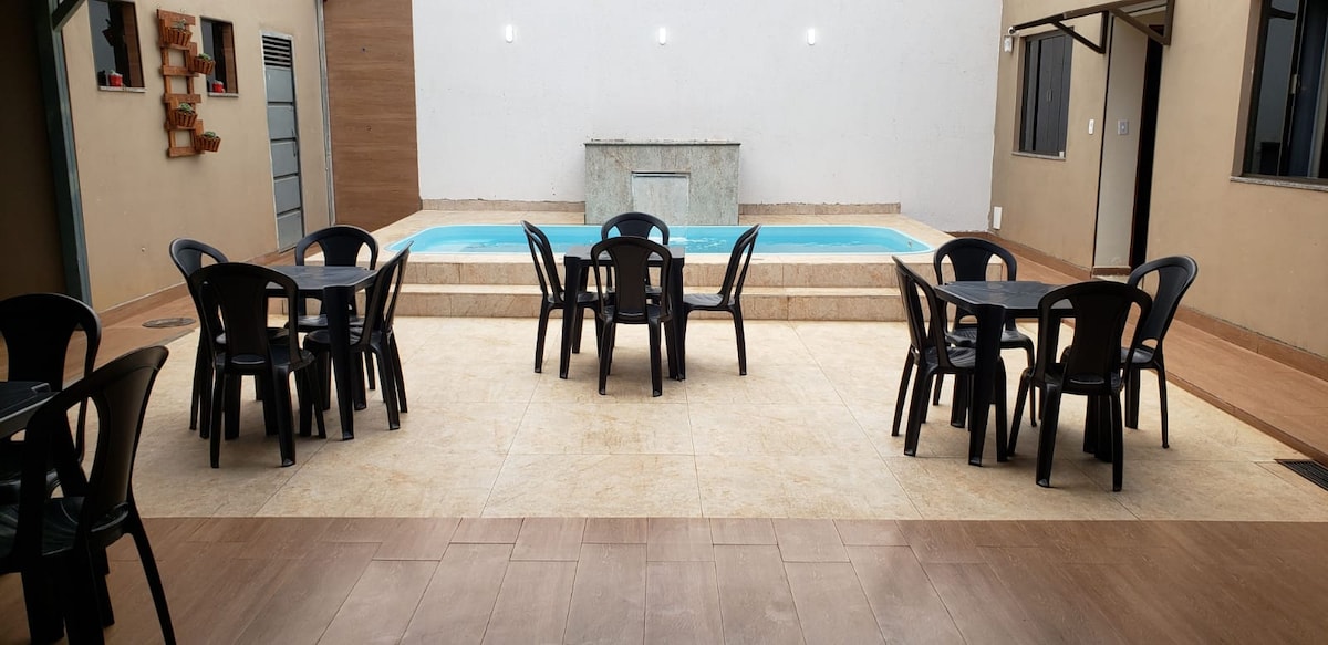 An outdoor area features a rectangular pool surrounded by a tiled terrace. Black plastic tables and chairs are arranged in a shaded area, while the pool's stone backdrop provides a clean and modern look. Light fixtures are mounted on the wall, adding illumination.