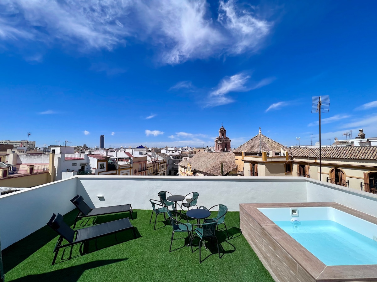 A rooftop terrace features a small pool surrounded by a wooden deck. Lounge chairs and a dining set with four chairs are positioned on lush green artificial grass. The skyline of Seville provides a scenic backdrop, showcasing traditional rooftops and historic structures under a clear blue sky.
