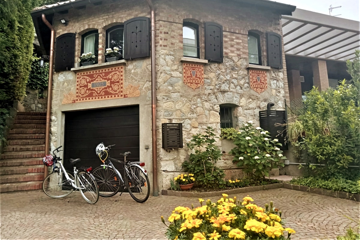 An inviting stone villa features a rustic façade with decorative brick patterns. Bicycles are parked in front of the garage, surrounded by neatly arranged flower beds. Two arched windows adorned with flower boxes add charm to the entrance, while a cobblestone pathway leads towards the entrance.