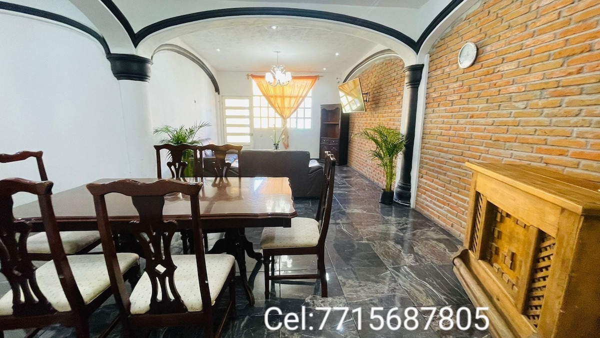 A spacious dining area is furnished with a wooden table and several matching chairs. A comfortable living space is visible, featuring a gray sofa and wall-mounted entertainment unit. The room is accented by natural light filtering through a window, with a decorative plant enhancing the ambiance.