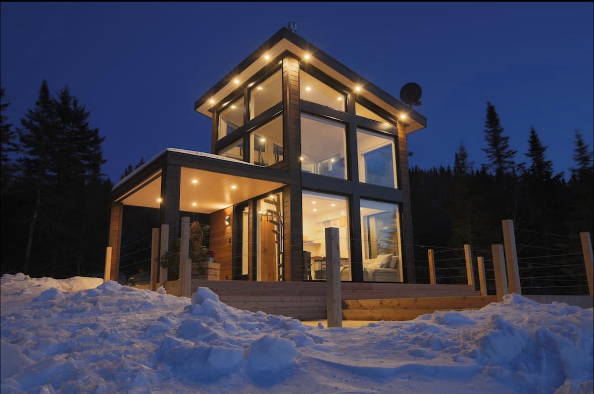 A modern chalet with extensive glass facades stands in a snowy landscape, illuminated by warm outdoor lights. The structure is surrounded by trees, with a wooden deck leading to the entrance. Snow blankets the ground, contributing to the serene winter setting.