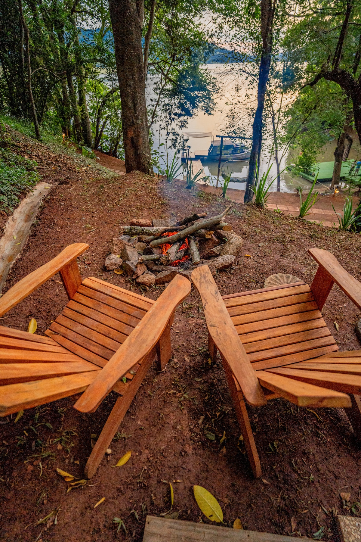 Two wooden chairs are positioned around a fire pit, offering a cozy spot for gatherings. A tranquil view of the water and nearby trees is visible in the background, enhancing the connection to nature.
