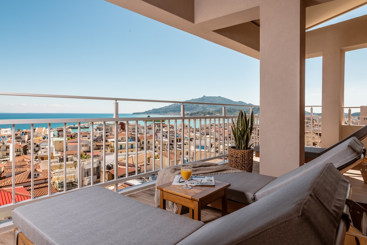 A spacious balcony area features two sun loungers and a small side table, inviting relaxation. Scenic views of the sea and Zante town are visible, with green hills in the distance, providing a serene backdrop for outdoor enjoyment.