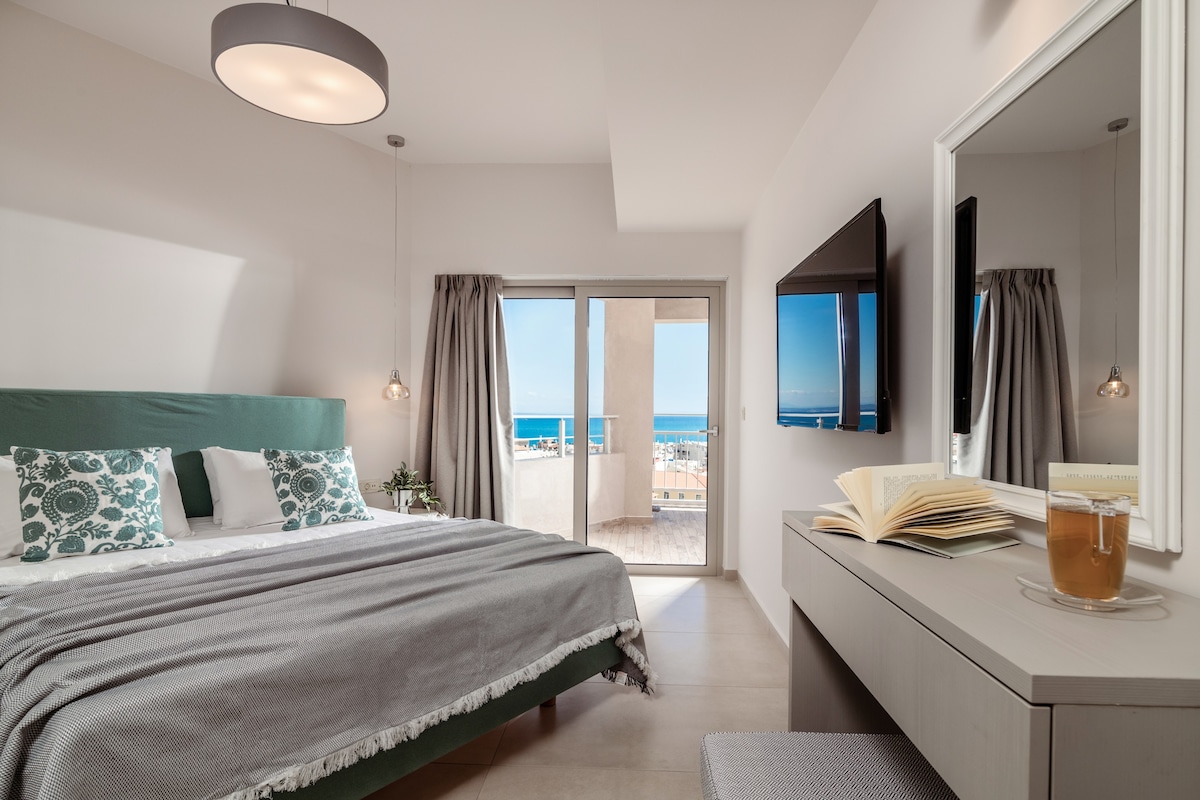 A bright bedroom is presented with a spacious bed featuring green accents, complemented by leafy patterned pillows. A large window allows natural light and offers views of the sea. A cozy reading area is visible with a small table, book, and a beverage, enhancing the serene ambiance.