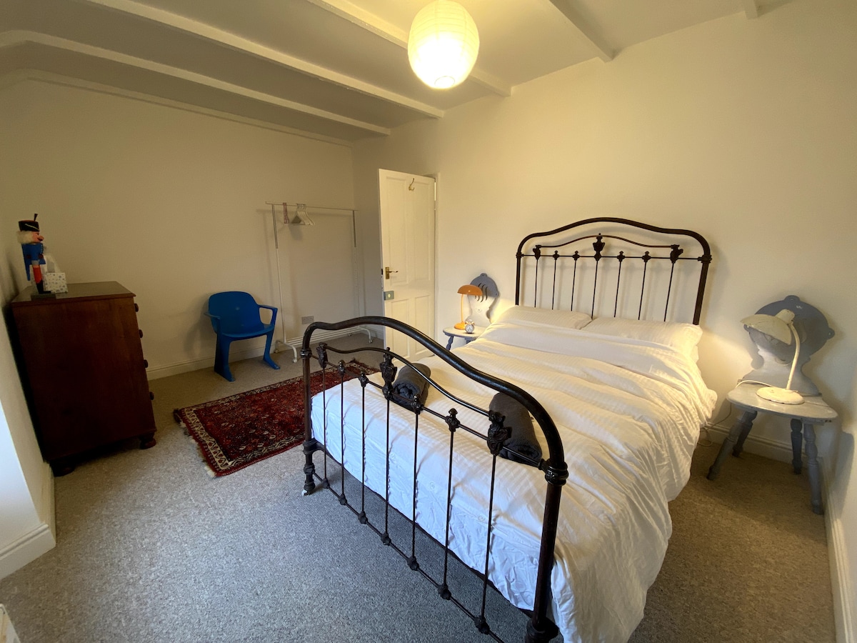A bright bedroom features a double metal bed with crisp white bedding. A wooden dresser and a small blue chair are positioned nearby, while a light fixture hangs above. The space is lightly decorated with a patterned rug on the carpeted floor.