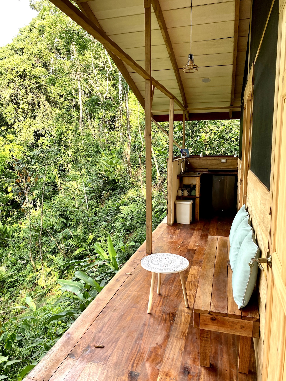 The outdoor terrace features a small table and two cushioned seats, inviting relaxation amidst the surrounding lush greenery. A kitchenette is visible, blending with the natural setting. Warm wooden flooring complements the cabin's rustic charm, with views of the vibrant jungle foliage in the background.