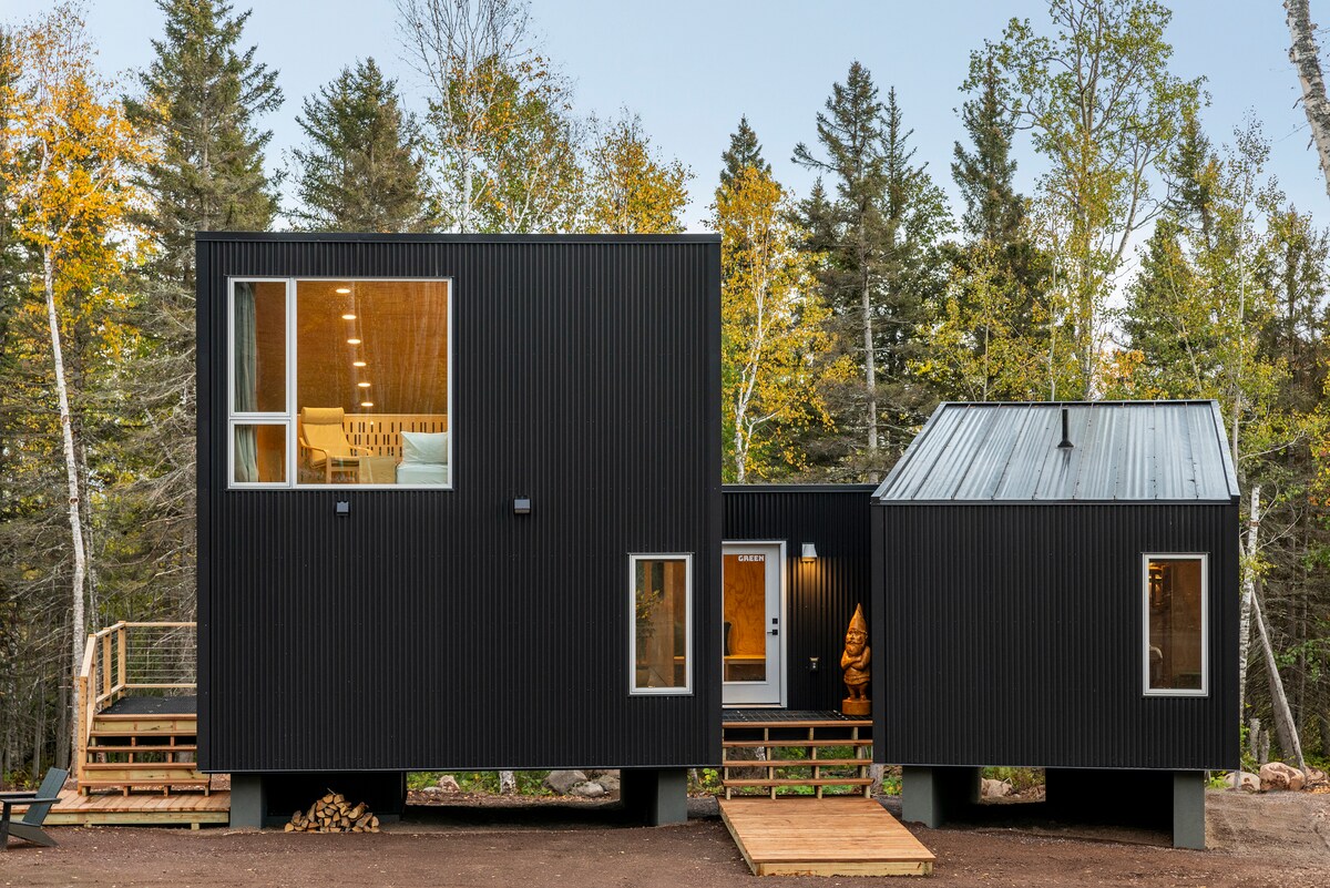 A modern, Scandinavian-style cabin is shown, featuring a sleek black exterior with multiple windows. The surrounding foliage displays autumn colors, and a wooden deck leads to the entrance. Elevated structure enhances connection with the natural environment.