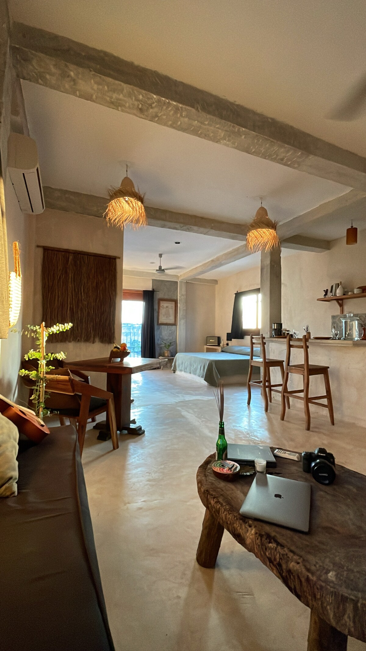 An inviting studio space features a cozy seating area with a dark sofa and a rustic wooden coffee table. Natural light enters through a window, illuminating a sleek kitchenette and a dining table. Decorative pendant lights hang from the ceiling, enhancing the overall ambiance.