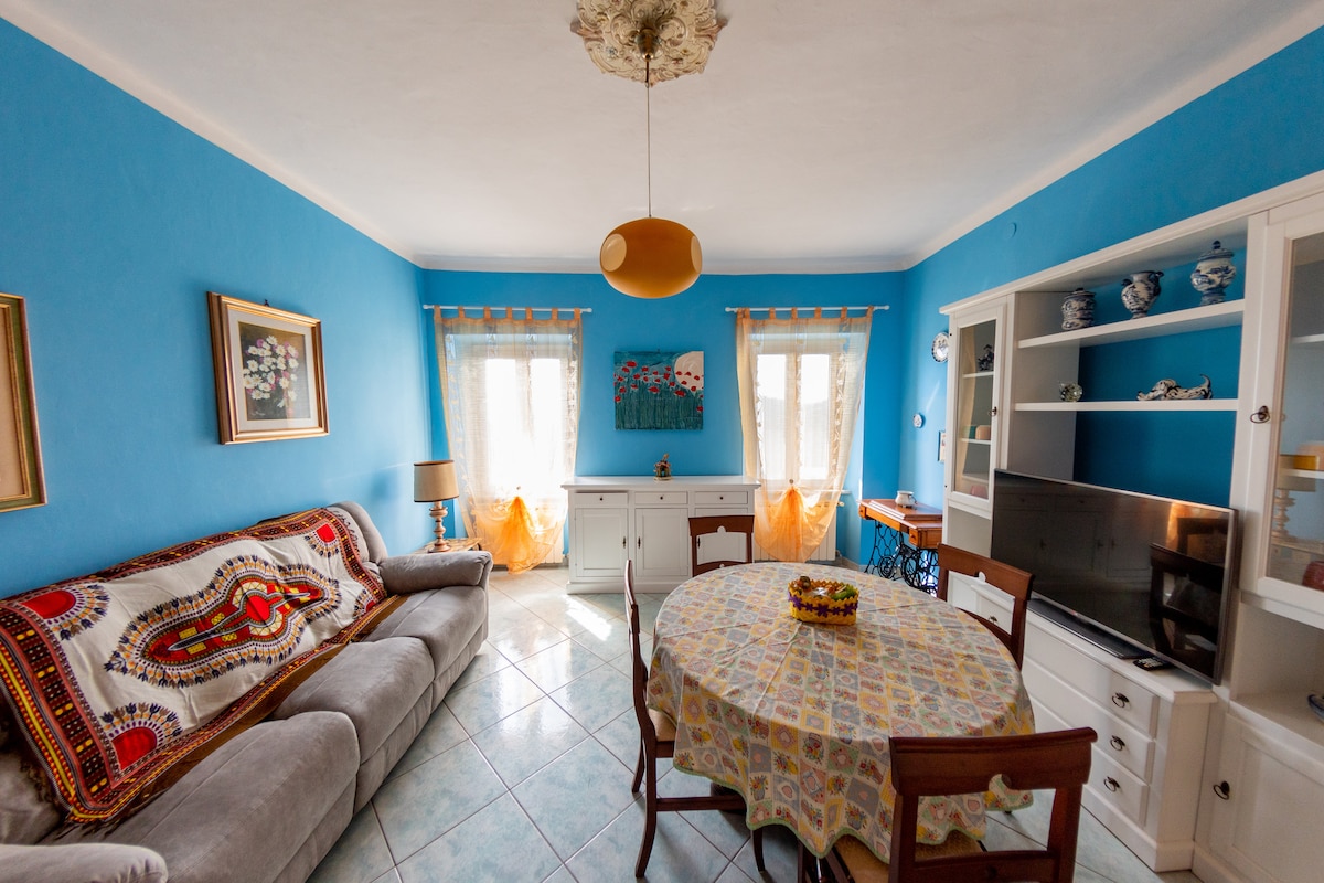 A spacious living area is highlighted by vibrant blue walls and large windows allowing natural light. A round table is set for dining, surrounded by wooden chairs. A comfortable sofa is adorned with a patterned throw, and a television sits within a light wood cabinet.