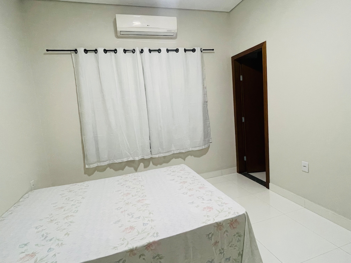A neutral-toned bedroom features a spacious bed with a floral-patterned coverlet. Sheer white curtains frame a window, allowing soft light to enter. An air conditioning unit is mounted above, and a door can be seen leading to another area of the home.