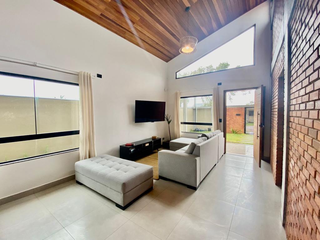 A modern living area is highlighted by wooden ceiling beams and large windows that allow natural light to fill the space. Comfortable seating is provided by a light gray sofa and an ottoman. A television is mounted on the wall opposite the seating arrangement.
