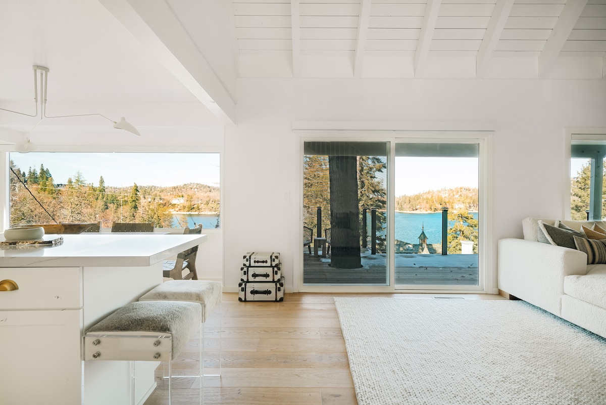 A light-filled living space features a spacious white couch and a large area rug. A kitchen island with seating is visible, while large glass doors offer views of the lake and surrounding trees, creating a connection to the outdoor deck.