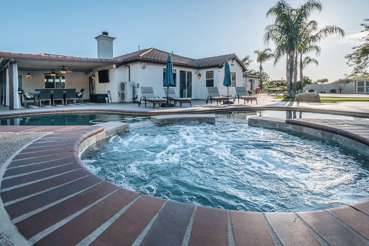 A hot tub is featured alongside a sparkling pool, surrounded by lounge chairs under shaded umbrellas. The spacious backyard includes palm trees and a glimpse of the home's exterior, highlighting a welcoming atmosphere for relaxation and socializing.
