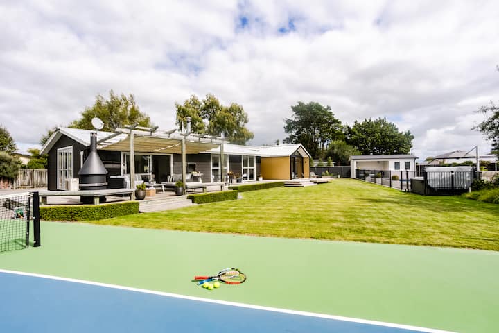 Spacious backyard with a modern house, covered patio, and a tennis court, surrounded by a well-maintained lawn and trees