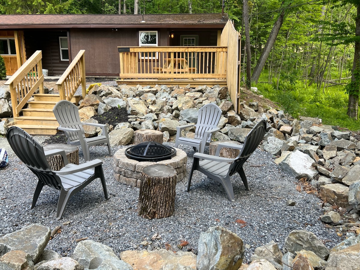 An outdoor fire pit area is surrounded by grey chairs positioned on a gravel surface. Stone pathways lead to the cabin in the background, which features a wooden deck and steps for easy access.