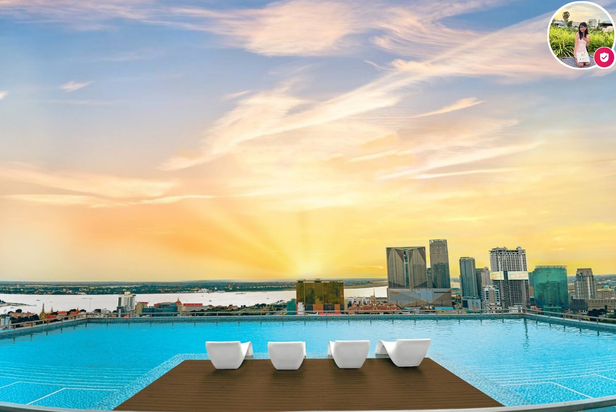 A rooftop pool is framed by a wooden deck, featuring three modern white chairs positioned for relaxation. The skyline is illuminated by a vibrant sunset, casting warm hues over the city and river below.