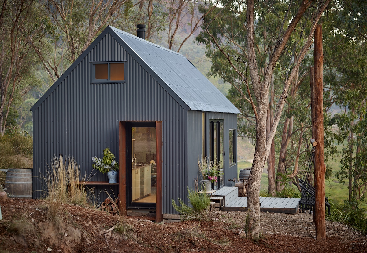 A modern off-grid cabin is set within a natural landscape, featuring a sleek grey exterior and large windows. The outdoor deck is surrounded by trees, and potted plants are placed near the entrance, inviting a connection with the serene environment.