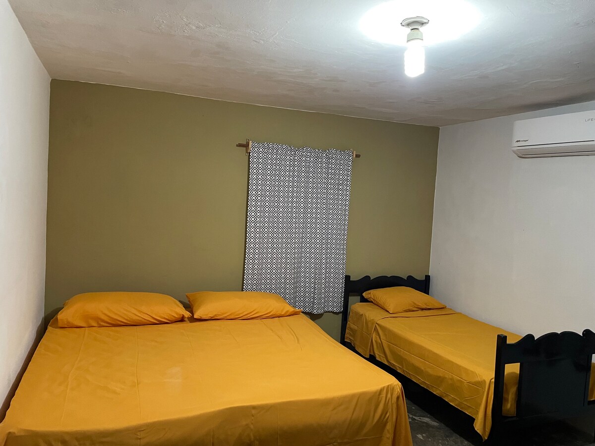A simple sleeping area is furnished with two beds, each dressed in yellow bedding. A window with patterned curtains provides natural light. An air conditioning unit is mounted on the wall, offering comfort during warm days.