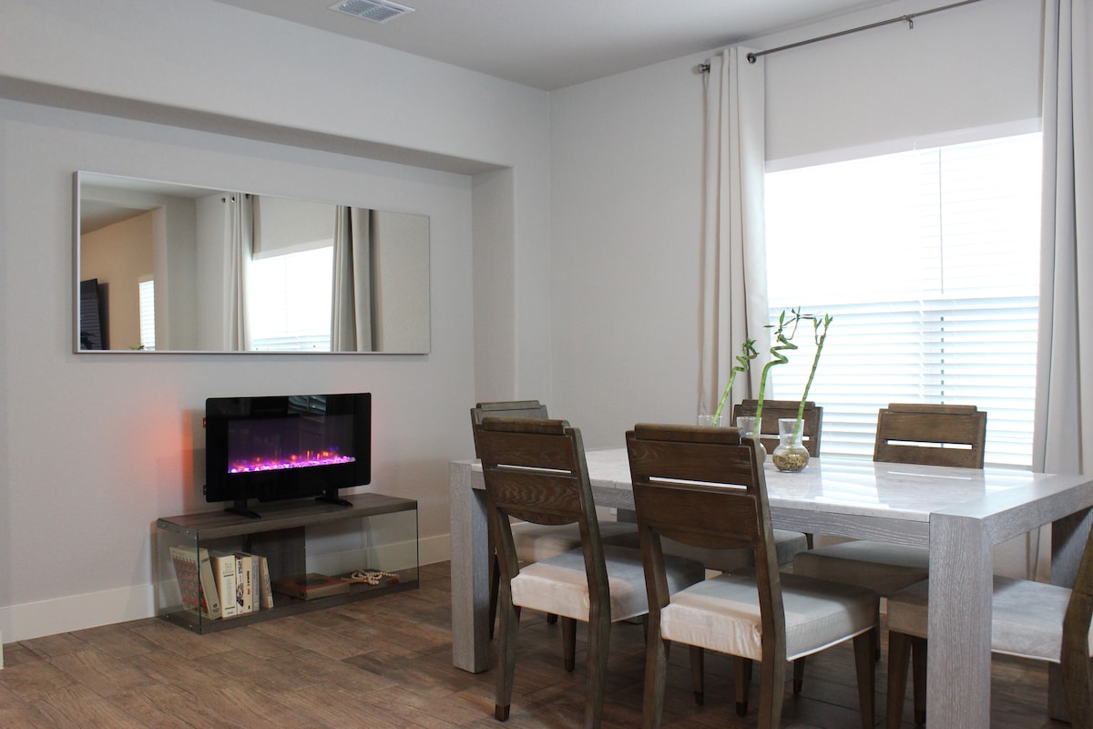 A modern dining area is featured with a rectangular table surrounded by six wooden chairs. A contemporary electric fireplace is positioned beneath a large mirror, while natural light filters through a window adorned with light curtains.