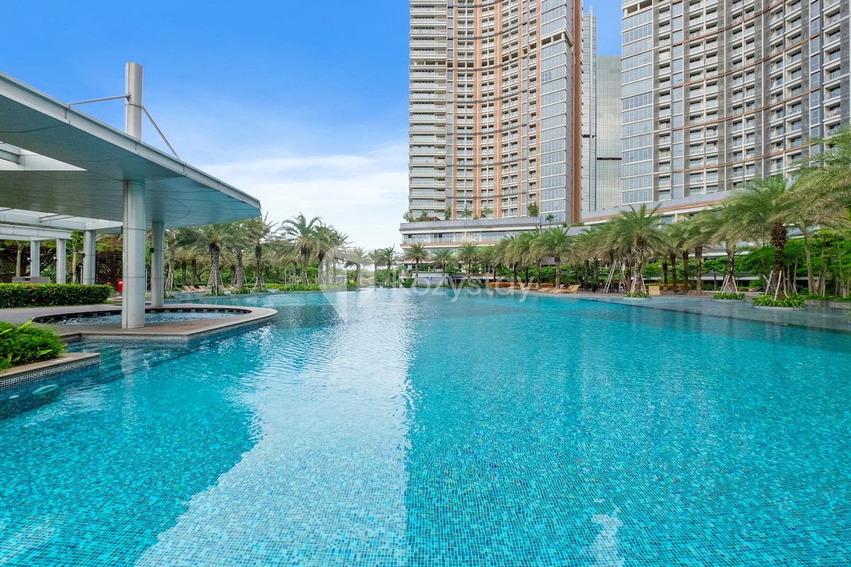 An expansive outdoor swimming pool is showcased, featuring crystal-clear turquoise water. Lush palm trees are visible along the poolside, with tall buildings in the background. A shaded area is provided, enhancing relaxation opportunities for guests.