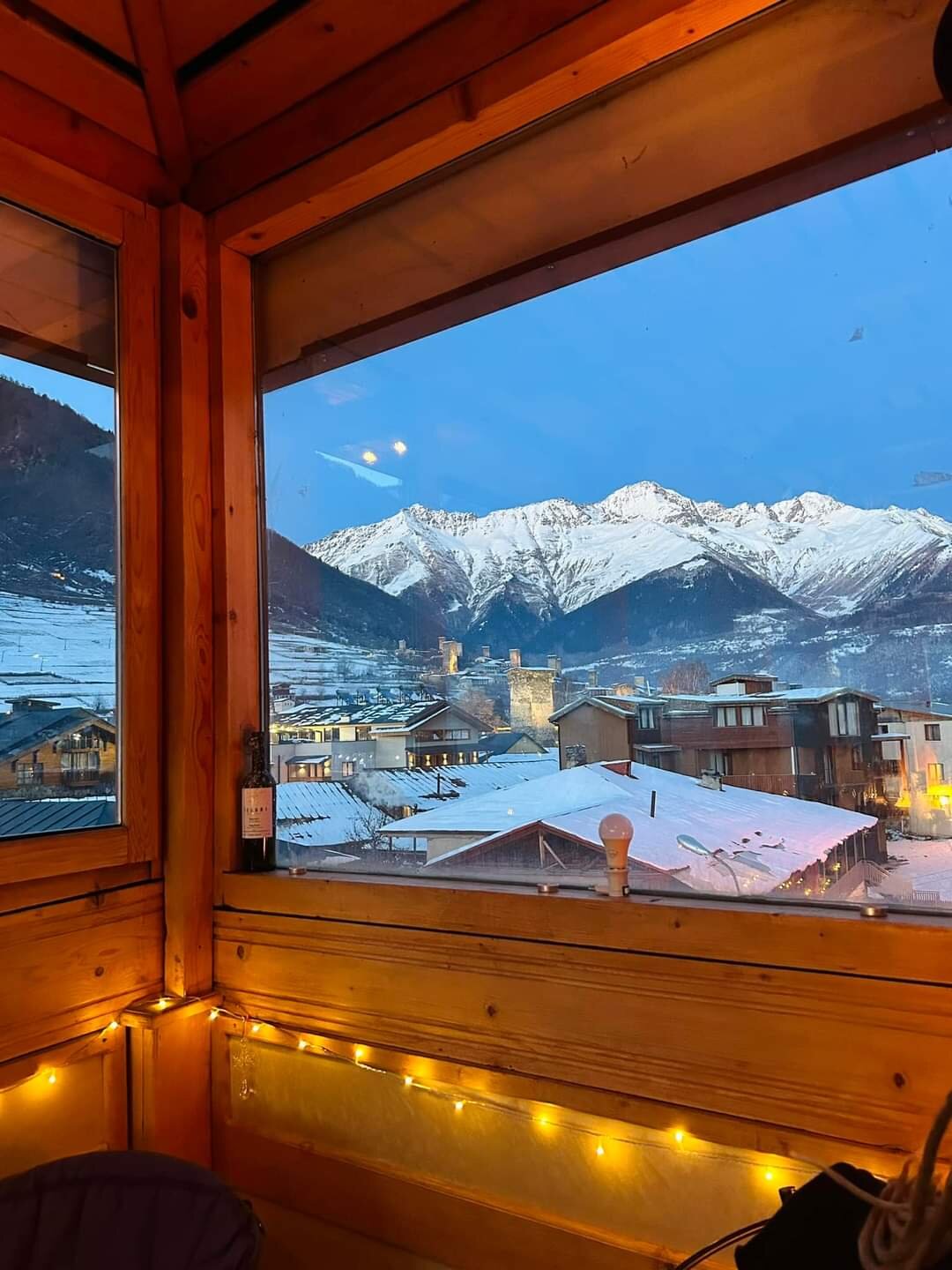 A panoramic view of snow-capped mountains is framed by large wooden windows, showcasing the serene landscape at dusk. Nearby roofs are blanketed in snow, and soft lighting is visible within the space, adding a cozy ambiance to the setting.