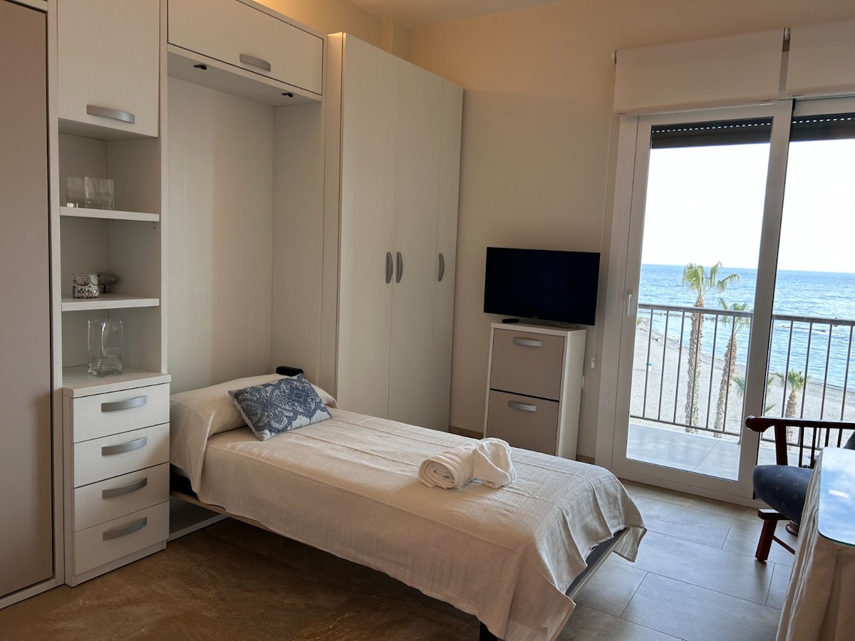A well-organized sleeping area features a single bed with a light-colored blanket and a decorative pillow. Storage cabinets and shelves are neatly integrated into the wall design. A flat-screen TV and a side table enhance convenience, while a glass door provides access to a balcony with a view of the sea.