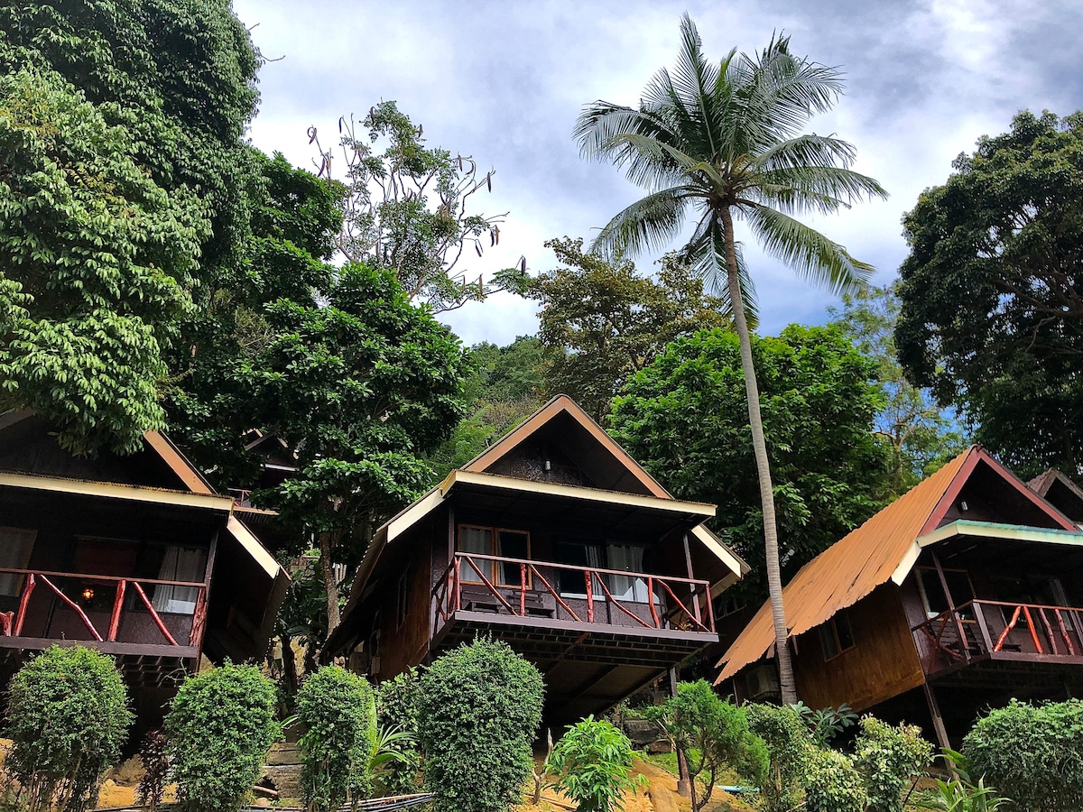 Rustic bungalows are nestled among lush tropical greenery, with wooden exteriors and balconies. A tall palm tree rises alongside the structures, complementing the natural setting. The hillside location is visible, indicating a slight upward slope from the ground.