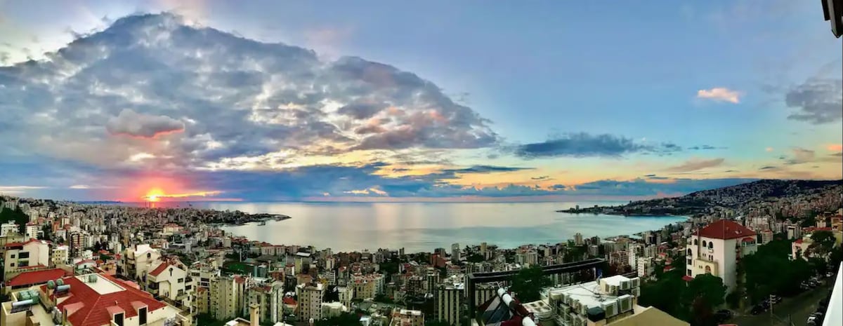 A panoramic view captures the shoreline of the Mediterranean Sea at sunset, with soft hues of orange and pink blending into a blue sky. The coastal city, characterized by a mix of buildings and greenery, stretches along the coast, showcasing a tranquil atmosphere.