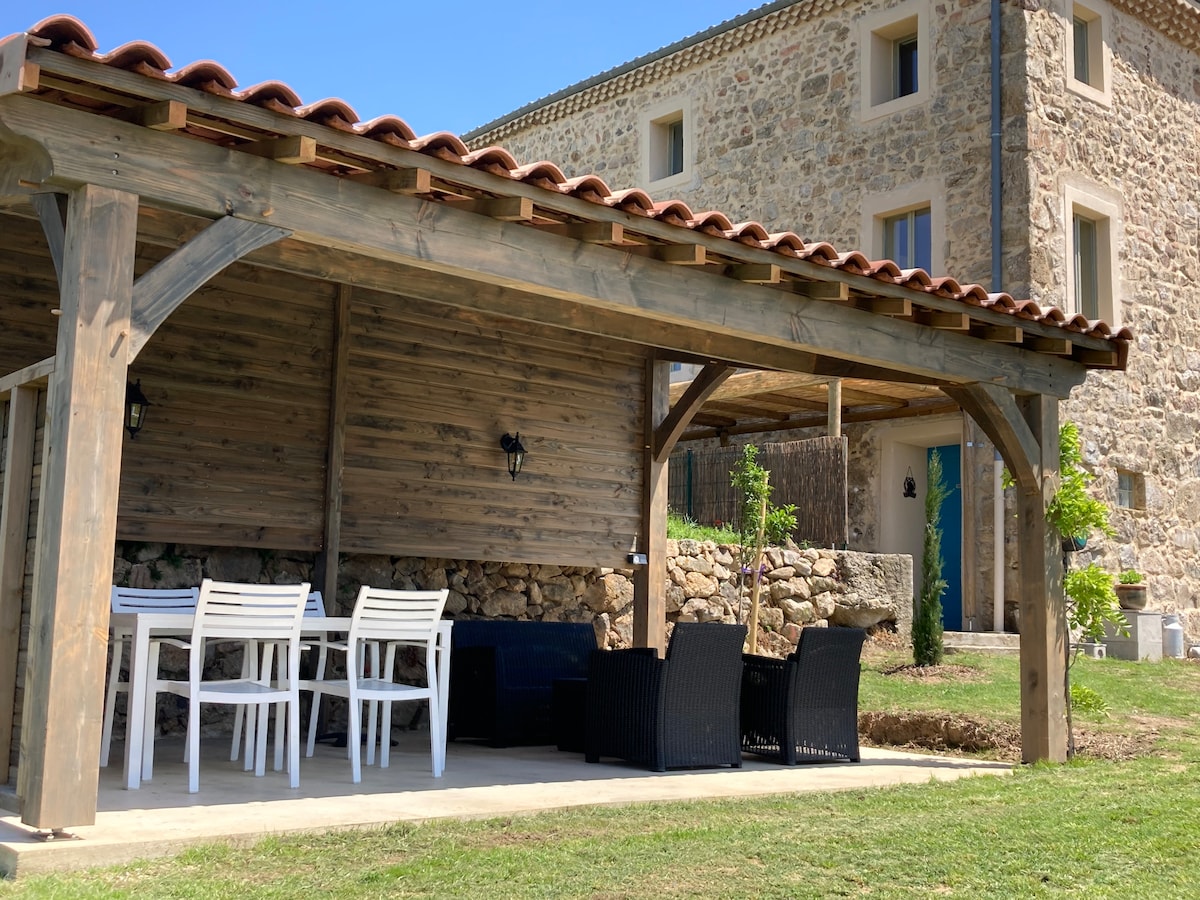 An outdoor seating area is featured under a wooden pergola, equipped with a table and white chairs. Two dark wicker chairs are positioned nearby. A stone building is visible in the background, showcasing a rustic facade and blue door, framed by clear blue skies.