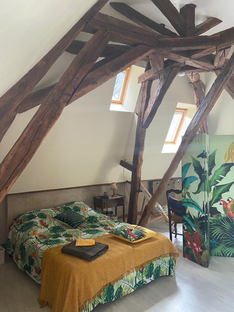 A cozy sleeping area features a bed adorned with a vibrant tropical-patterned duvet. Natural light filters through angled windows, highlighting exposed wooden beams that add character. A small bedside table and a foldable screen with lush green foliage complete the inviting space.
