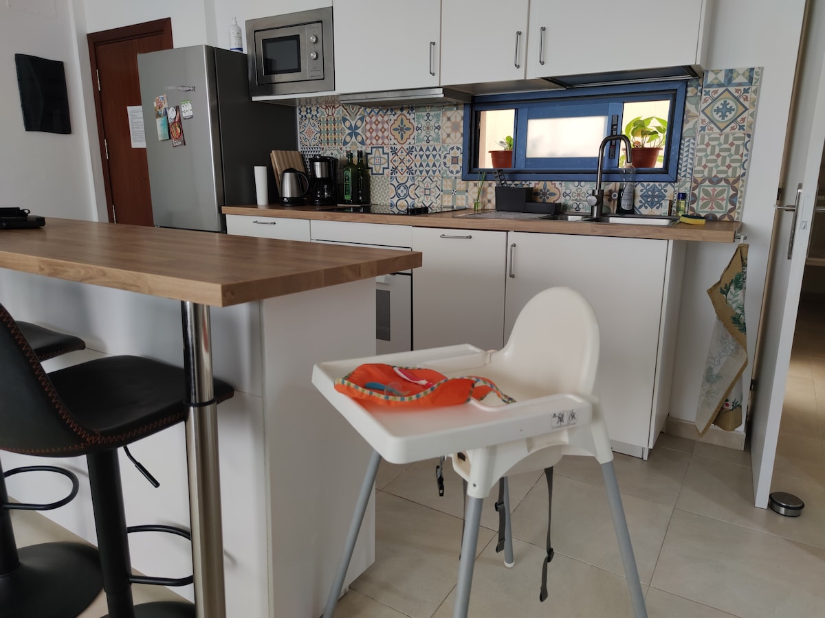 An open kitchen area is displayed, featuring modern appliances, including a microwave and coffee maker. Colorful tiles add visual interest to the backsplash. A high chair is positioned beside a bar with tall stools, creating a family-friendly atmosphere.