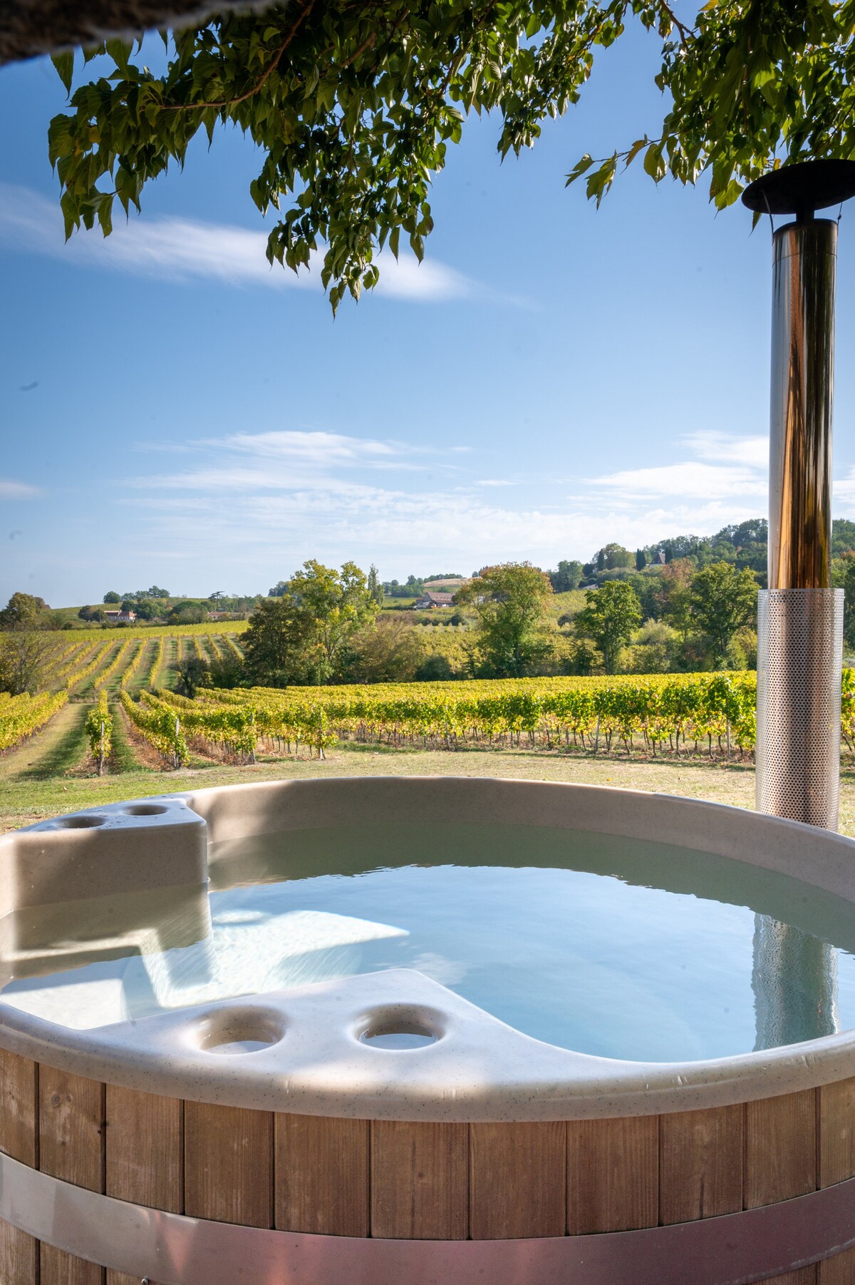 A wooden hot tub is seen on a terrace, surrounded by a picturesque vineyard landscape. Lush green hills and rows of grapevines stretch into the distance under a clear blue sky, creating a serene outdoor setting.