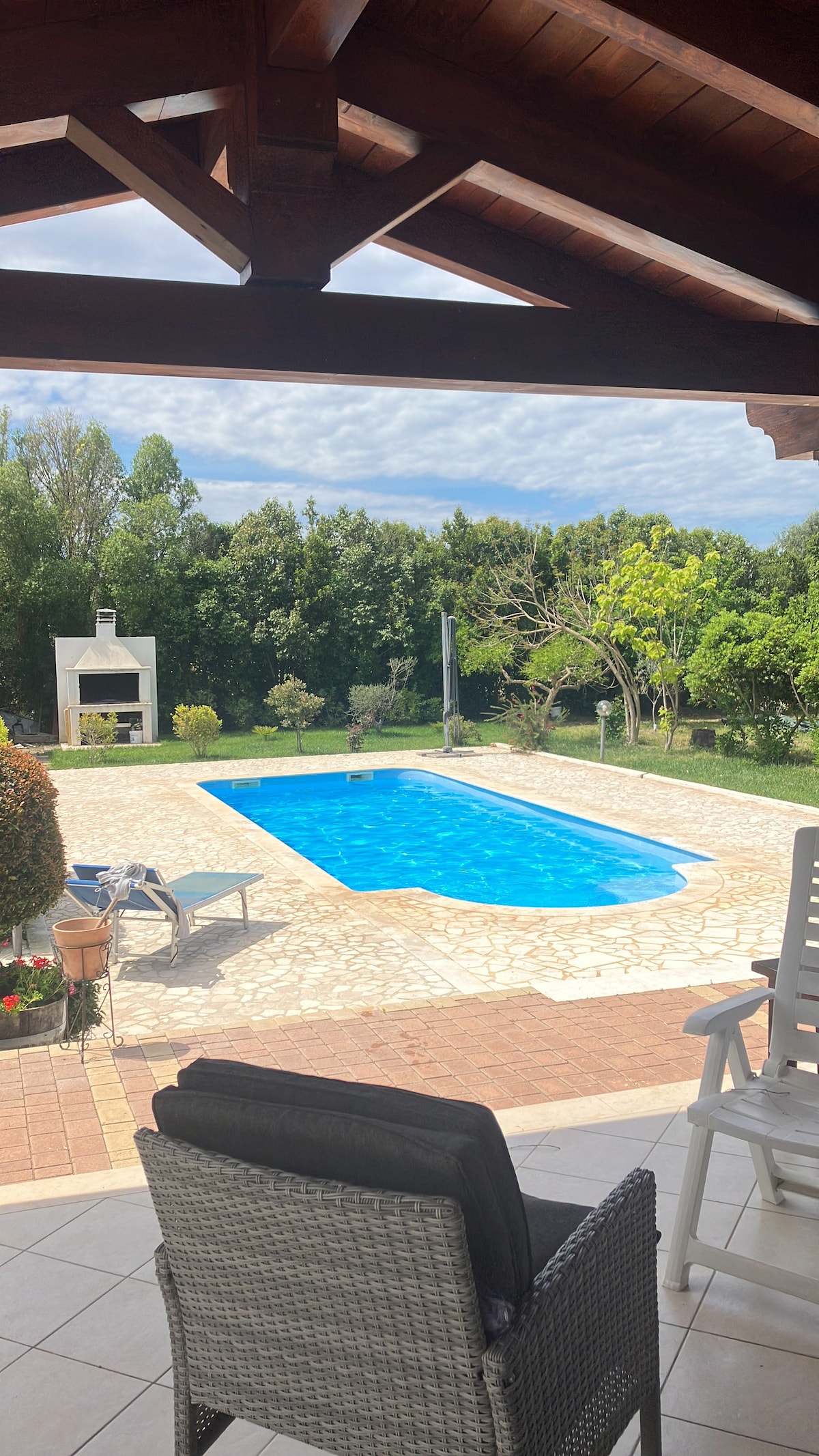 A tranquil outdoor space features a bright blue swimming pool surrounded by lush greenery. A patio area with lounge chairs and a barbecue provides comfortable seating, while a covered terrace offers shade. The scene is peaceful, emphasizing the relaxing countryside environment.