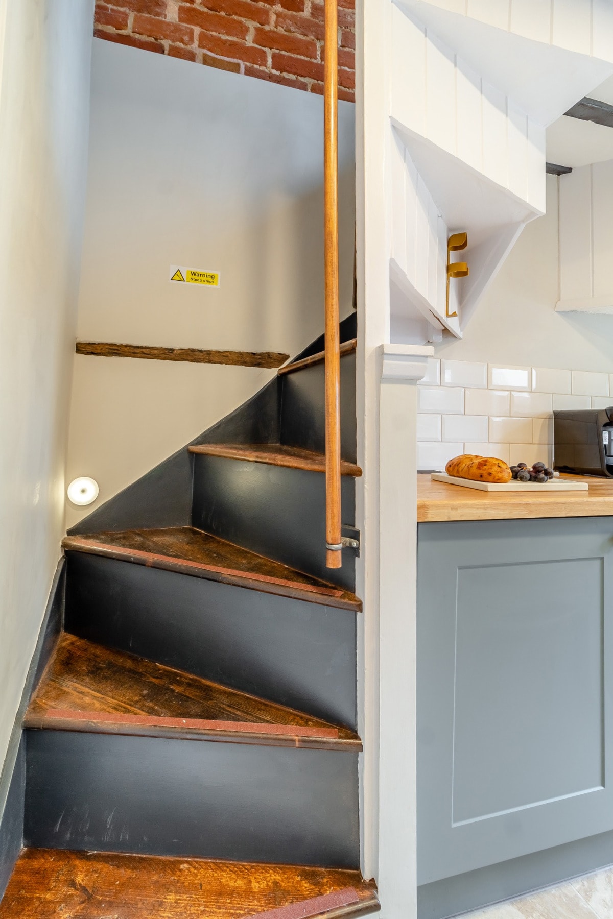 A spiral staircase with dark wooden steps leads upward, framed by brick and white wall surfaces. A smooth handrail is present for support, guiding the ascent to the upper level of the cottage.