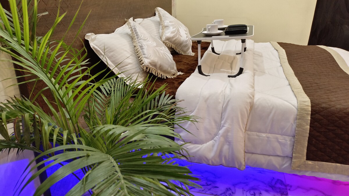 A neatly arranged sleeping area features a spacious bed with decorative pillows and a brown quilt. A small, portable table holds cups and a notepad beside the bed. Soft blue lighting highlights a decorative plant, creating a serene atmosphere.