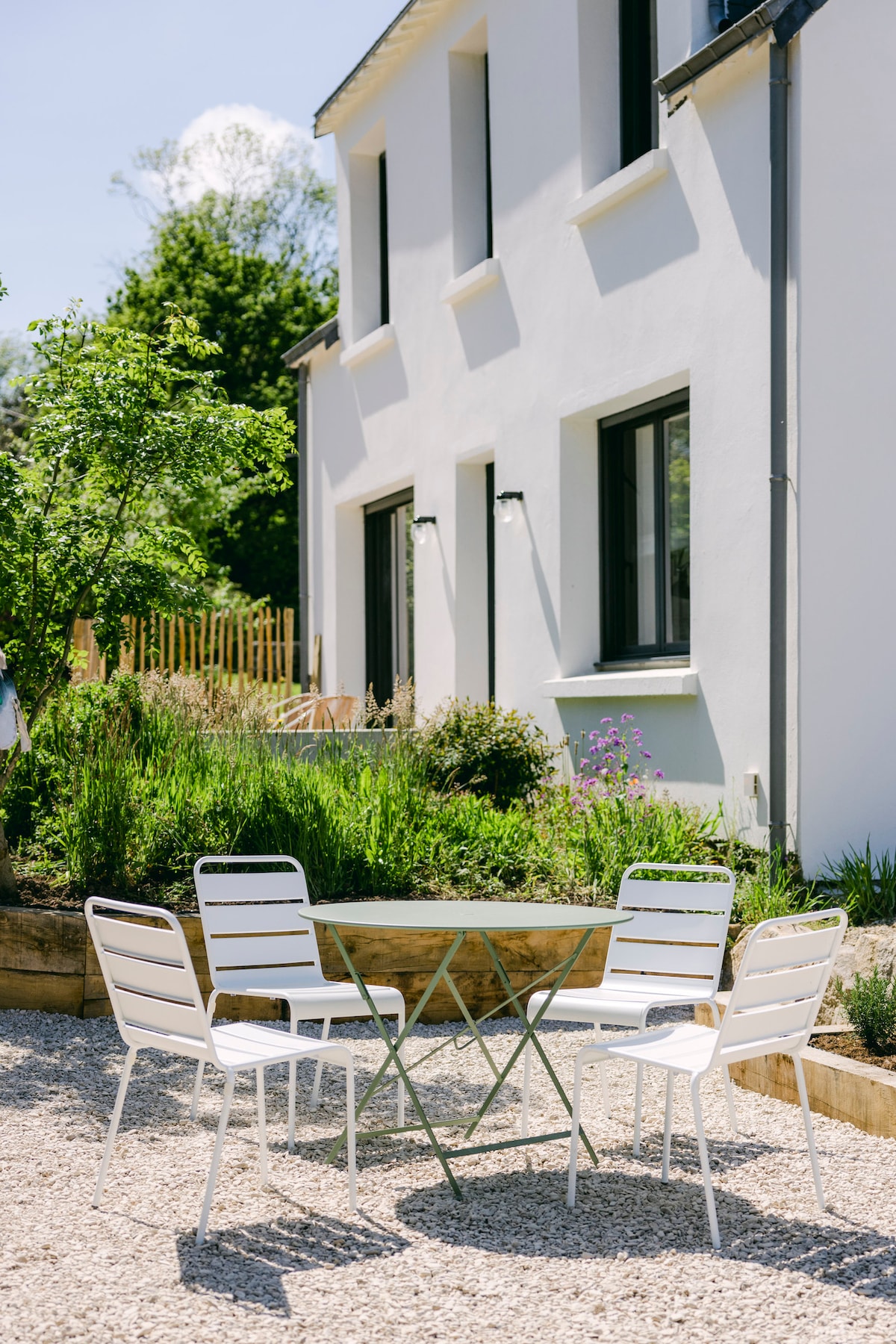 A serene outdoor area features a light-colored gravel surface with a round metal table surrounded by four white chairs. Lush greenery and blooming flowers provide a natural backdrop to the modern building's exterior.