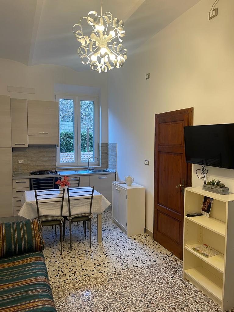 The living area features a sleek kitchen setup with modern cabinetry and a dining table set for four. A sofa adds comfort, while a wall-mounted TV is positioned nearby. Natural light filters through the large window, illuminating the elegant tiled floor.