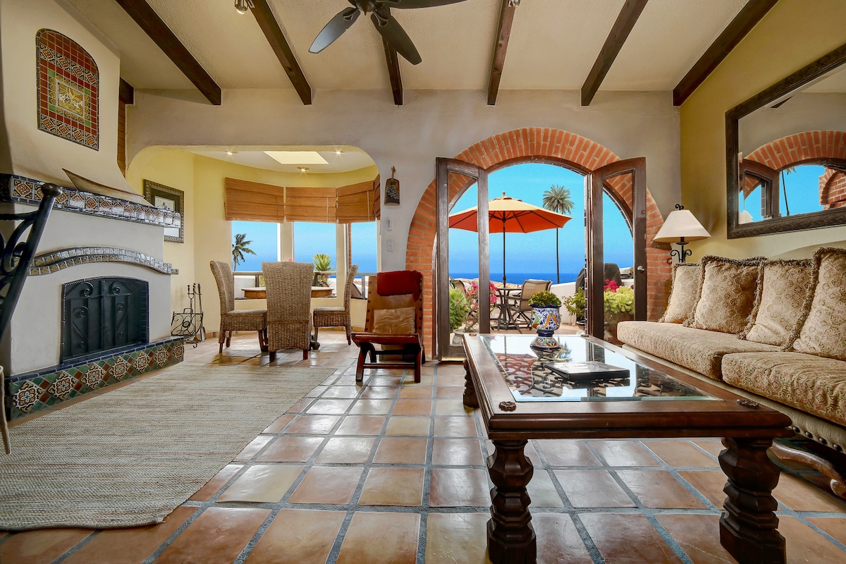 The inviting living area showcases Saltillo tile flooring and beams overhead. A coffee table sits in the center, surrounded by plush seating. Arched doors open to reveal picturesque ocean views and a sunny patio, highlighted by a vibrant umbrella and swaying palm trees.