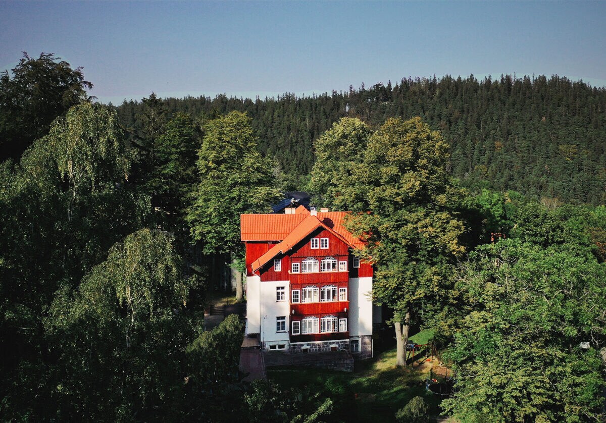 The charming villa, built in 1900, is surrounded by lush greenery and tall trees. Its striking red roof and white walls provide a classic contrast against the landscape. Large windows are visible, enhancing the connection with the natural environment.